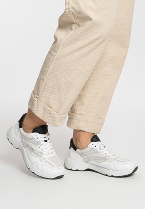 Persona che indossa pantaloni in velluto beige e scarpe da ginnastica bianche con accenti neri, in piedi su una superficie bianca uniforme.