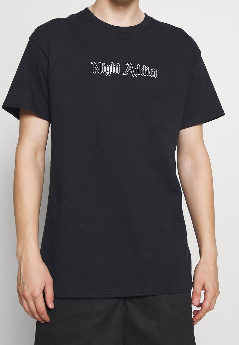 Černé bavlněné tričko s krátkými rukávy a bílou výšivkou s nápisem „Night Addict“ ve stylizovaném písmu přes hrudník.