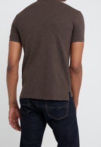 Man som bär en brun kortärmad polo och mörkblå jeans, sedd bakifrån mot en enkel ljus bakgrund.