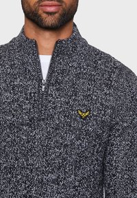 Strikket zip-sweater i mørkegrå med lysegrå stænk. Har en stående krave og et guldemblem på brystet. Blød tekstur.