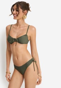 Μαγιό μπικίνι σε ελιά πράσινο χρώμα με ελαφρώς επενδυμένο τοπ που έχει δεμένο κόμπο στο κέντρο και ρυθμιζόμενους ιμάντες· ταιριαστά σλιπ με χαμηλή μέση και πλευρικές κορδέλες για δέσιμο.
