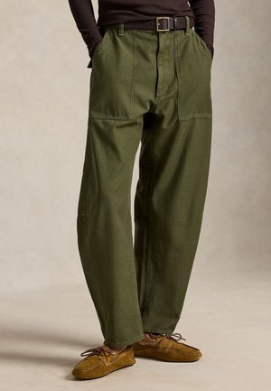 Homme portant un pantalon ample vert olive avec de grandes poches avant, une ceinture noire, des chaussures marron style mocassins, et une chemise à manches longues foncée, les mains dans les poches.