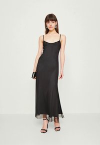Black maxi dress with thin straps, flowing silhouette, and sheer hem. Smooth fabric with a sleek texture. Paired with black strappy heels.