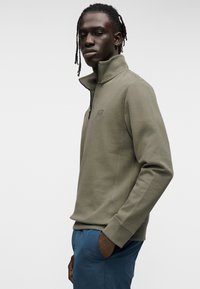 Πράσινο-ελιάς φούτερ με φερμουάρ μέχρι το στήθος, με ψηλό γιακά και μικρό λογότυπο στο στήθος. Μαλακό ύφασμα, εφαρμοστή σχεδίαση και ριμπ μανσέτες.