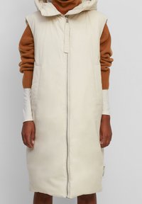 Gilet en duvet long couleur crème avec fermeture éclair sur le devant, grande capuche et poches latérales. Tissu texturé avec une finition brillante et détail d'étiquette sur le côté.