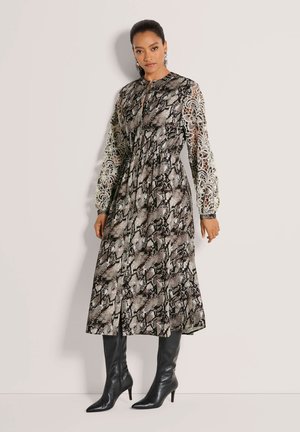 Frau trägt ein Midikleid mit Schlangenprint, florale Spitzenärmel und schwarze kniehohe Stiefel mit Absatz, steht vor einer schlichten Wand.