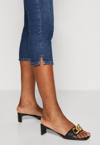 Svarta slidesandaler med en liten blockklack, med en guldkedja som accent på remmen. Bärs med avklippta blå denimjeans.