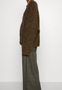 Brauner Strickcardigan mit strukturiertem Finish, langen Ärmeln und zwei vorderen Taschen, kombiniert mit lockeren olivgrünen Hosen.