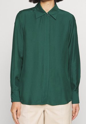 Osoba ubrana w ciemnozieloną koszulę na długi rękaw zapinaną na guziki oraz beżowe spodnie, stojąca z rozluźnionymi rękami wzdłuż ciała na jednolitym tle.