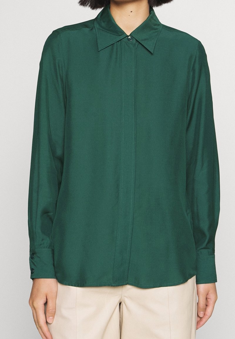 Personne portant une chemise boutonnée à manches longues vert foncé avec un pantalon beige, debout, bras détendus le long du corps, devant un fond uni.