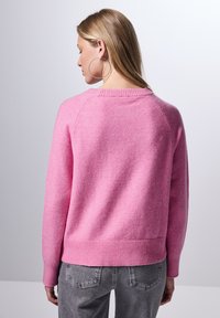 Pull en rose en matière douce et texturée avec un col rond et un ourlet côtelé. Il présente des manches raglan et une coupe décontractée, associé à un jean gris.