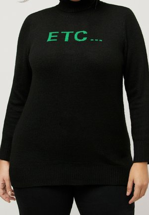 Personne portant un pull à col roulé noir avec un texte vert "ETC..." sur la poitrine, debout devant un fond clair uni.