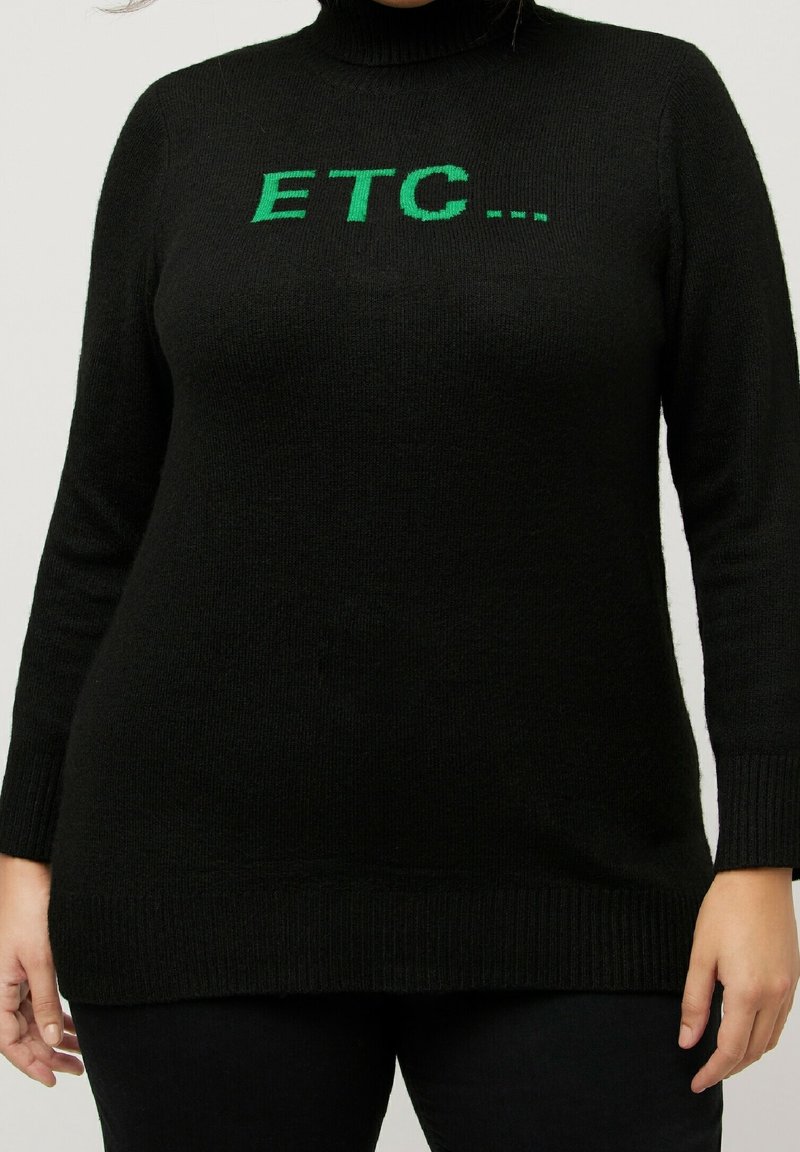 Personne portant un pull à col roulé noir avec un texte vert "ETC..." sur la poitrine, debout devant un fond clair uni.
