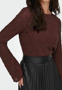 Donna che indossa un top in pizzo floreale marrone scuro a maniche lunghe, infilato in una gonna nera plissettata a vita alta con cintura larga.