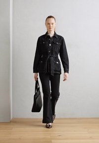 Veste en denim noire avec poches avant et ceinture, associée à un pantalon évasé noir et des chaussures à motif. Le mannequin porte un sac tote noir.