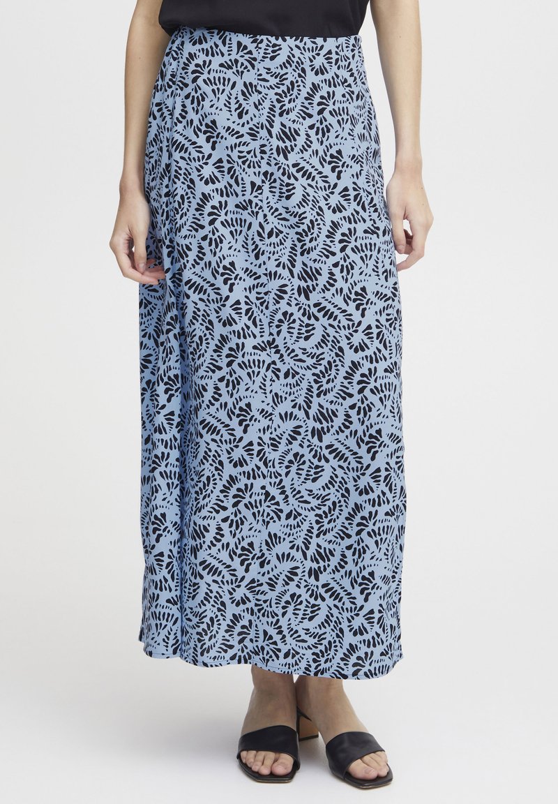 Falda maxi en azul claro con un estampado de hojas negras. Tejido fluido, cintura alta y detalle de abertura lateral. Combinada con sandalias de tacón negras.