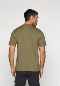 Maglietta a maniche corte verde oliva con scollatura a girocollo. Tessuto di cotone morbido, forma rettangolare. Indossata con pantaloni neri con strisce bianche.