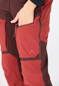 Pantalons d'extérieur rouges et bruns avec une poche zippée, un tissu durable, une forme structurée et un accent de logo au niveau de la cuisse.