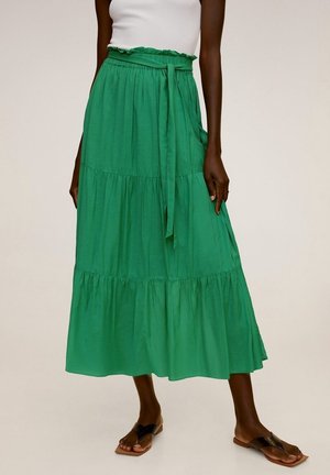 Femme portant une jupe midi verte à volants avec une ceinture nouée à la taille et des sandales plates noires, associée à un top blanc sans manches.