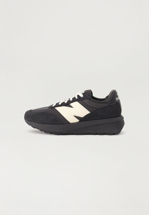 Schwarzer Sportschuh mit weißem "N"-Logo, weißen Schnürsenkeln und dicker schwarzer Sohle, von der Seite auf einem schlichten Hintergrund dargestellt.