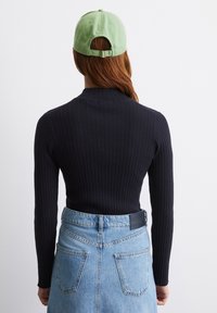 Person mit langen braunen Haaren, die eine hellgrüne Mütze verkehrt herum trägt, ein schwarzes, geripptes Langarmoberteil und einen hoch taillierten hellblauen Jeansrock.