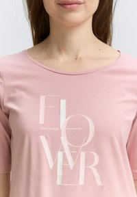 Helles rosa T-Shirt mit kurzen Ärmeln, das den Text "FLOWER" mit einem glänzenden Finish und kleinen Strasssteinen zeigt. Weicher, glatter Stoff.