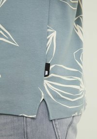 Détail de l'ourlet d'une chemise bleue avec un motif abstrait de feuilles blanches portée sur un jean en denim gris clair.