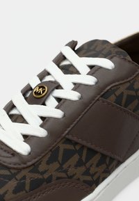 Rjavi vzorčasti športni copati z belimi vezalkami, s rjavim usnjenim poudarkom, zlatim logotipom in teksturirano površino.