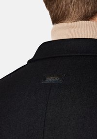 Czarny wełniany płaszcz o gładkiej fakturze z skórzaną metką na tylnym kołnierzu, zestawiony z beżowym golfem w prążki na dole.