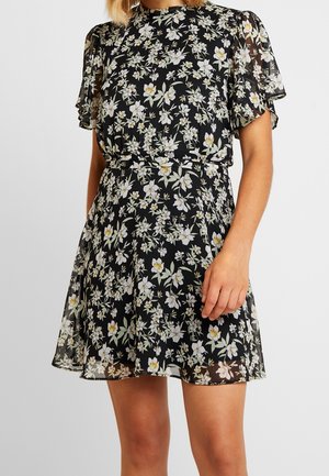 Kvinde iført en sort blomsterkjole med korte flagrende ærmer og en markeret talje, med hvide og gule blomsterprint.