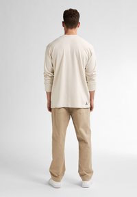 Lichtbeige langetermijn shirt met een ronde hals, gecombineerd met licht khaki broek. Het shirt heeft opgerolde mouwen en een klein logo aan de zoom.