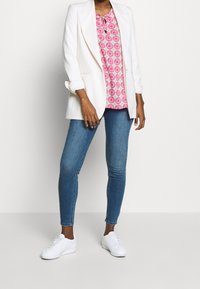 Blazer blanc porté sur un chemisier rose à motifs avec un col noué, associé à un jean skinny bleu et des baskets blanches. Textures lisses et coupe ajustée.