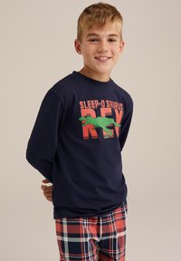 Mornarska majica z dolgimi rokavi z grafiko dinozavra in napisom "SLEEP-O SAURUS REX"; kombinirana s karirastimi hlačami rdeče, smetane in mornarske barve.