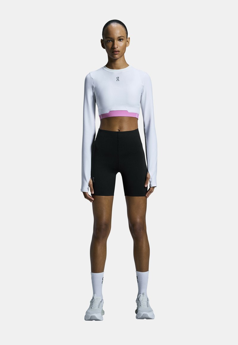 Weißes, langärmeliges, cropped Oberteil mit pinken Akzenten, kombiniert mit schwarzen High-Waist-Shorts. Das Model trägt weiße Socken und Laufschuhe.