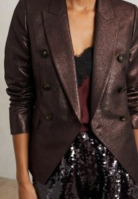 Dvouvrstvý sako v tmavě kovové fialové s lesklou texturou. Má zaoblené tmavé knoflíky a strukturované klopy, vrstvené přes krajkový top.