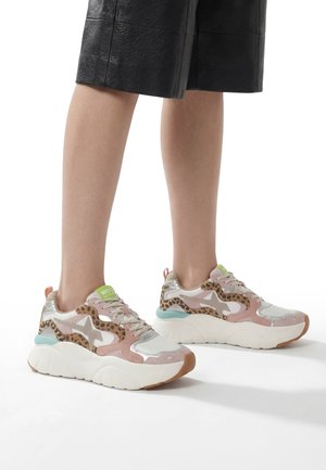 Ben iført sorte lædershorts og chunky sneakers med pink, hvide, leopardprint og grønne accenter på en hvid baggrund.