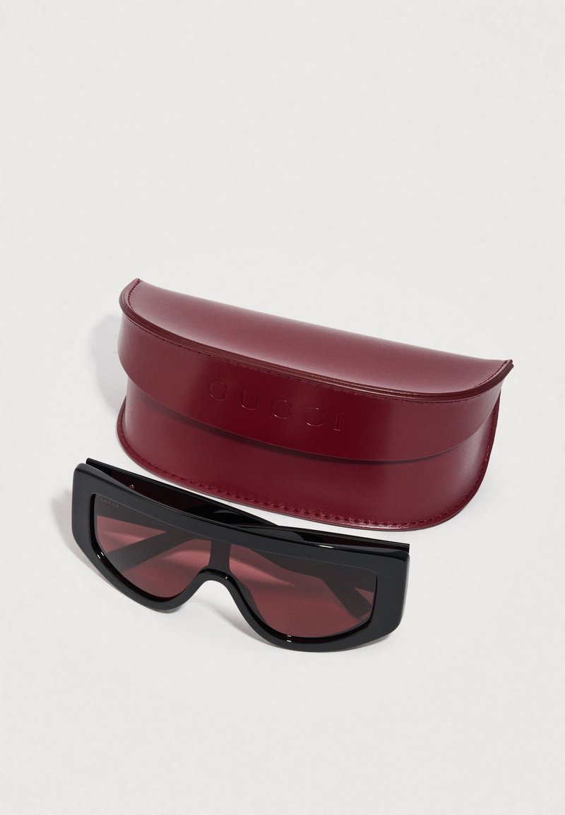 Black sunglasses with a unique angular design, paired with a burgundy leather case embossed with "GUCCI." Textured surface and smooth finish.