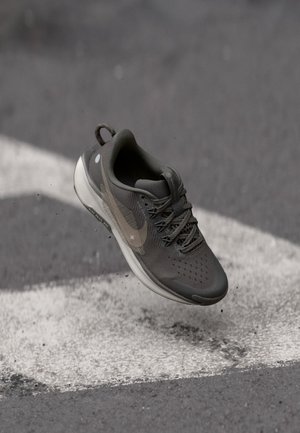 Grå sportskor med en texturerad ovandel, snörning och en vit sula. Har en subtil logotyp och reflexdetaljer. Dammkorn synliga.