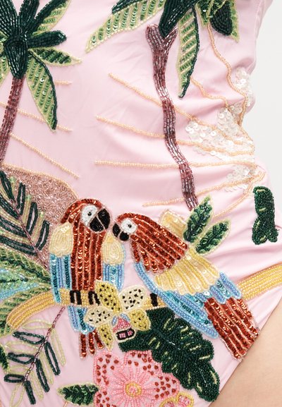 Deux perroquets en perles avec des plumes rouges, bleues et jaunes perchés sur une branche entourée de feuilles vertes et de fleurs roses sur un tissu rose.