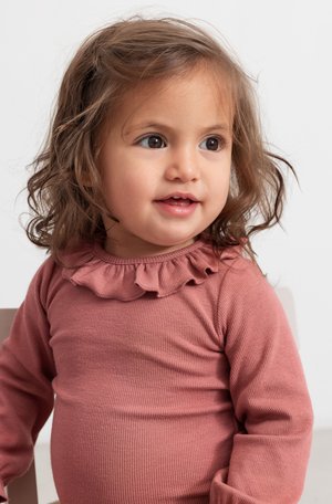 Top à manches longues en rose poussiéreux avec un col froncé. Texture douce, forme ajustée et encolure ronde. Enfant avec des cheveux ondulés.