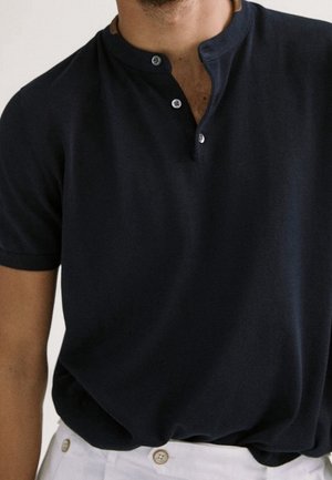 Homme portant un polo henley noir à manches courtes rentré dans un pantalon blanc boutonné, visible des épaules à la taille.