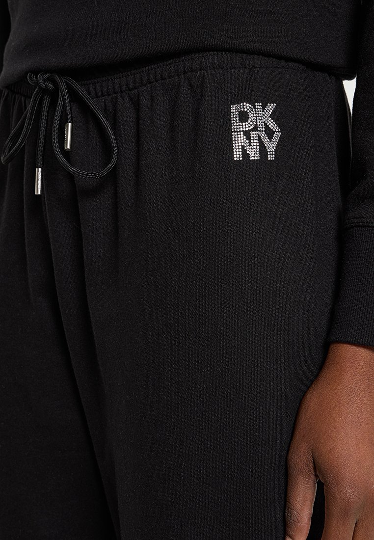 Zbliżenie na czarne spodnie dresowe z kieszenią na sznurek i logo "DKNY" w rhinestones na biodrze, noszone z pasującą czarną bluzą z długim rękawem.