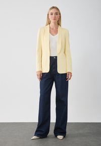 Blazer jaune ajusté avec des revers crantés sur un haut blanc à col en V, associé à un pantalon en denim bleu foncé à coupe ample et des chaussures pointues texturées.