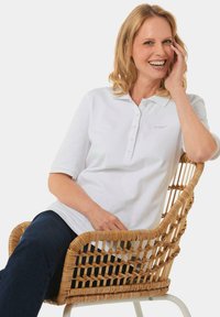Weisses Poloshirt mit kurzen Ärmeln, Knopfleiste und gesticktem Logo, kombiniert mit dunklen Jeans, während er/sie in einem geflochtenen Rattanstuhl sitzt.