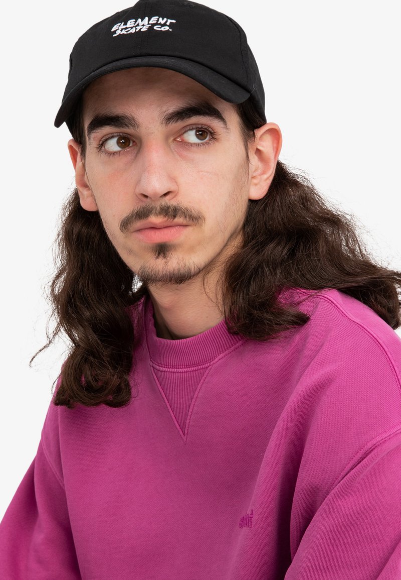 Un hombre lleva un sudadera rosa claro con un cuello redondo y un logo sutil, combinada con una gorra negra con texto en blanco.
