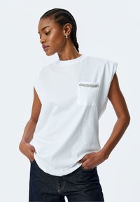 Hvid oversized ærmeløs t-shirt i bomuld med en brystlomme prydet med en sølvkæde-detalje. Glat tekstur, afslappet pasform.