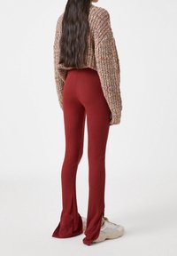 Donna in piedi con pantaloni aderenti color ruggine con spacchi, scarpe da ginnastica chunky beige e un maglione lavorato multicolore con texture, di spalle rispetto all'osservatore.