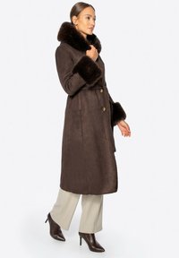 Brązowy płaszcz z sztucznego zamszu z futrzanym kołnierzem i mankietami, posiada dwa guziki oraz pasek w talii, zestawiony z jasnymi spodniami i szpilkami.