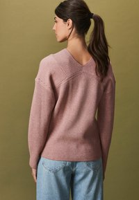 Helles rosa geripptes Sweatshirt mit weitem V-Ausschnitt und legeren Schultern. Kombiniert mit lockeren blauen Jeans. Der Hintergrund ist in sanftem Grün.