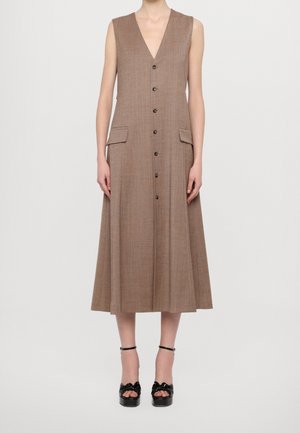 Brązowa sukienka midi bez rękawów z guzikami z przodu i kieszeniami z klapami, noszona z czarnymi sandałami na platformie z paskami wokół kostek.